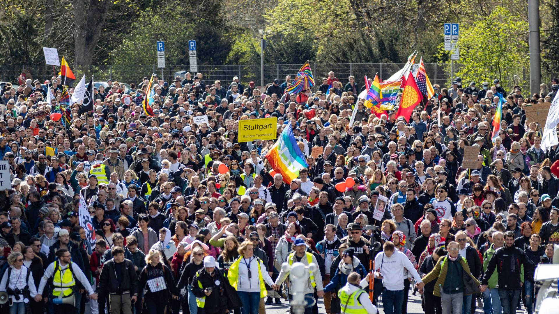 Eine Menschenmenge mit Plakaten auf einer Straße in Stuttgart. Menschen in neongelben Westen stehen vorn, einige Demonstranten schwenken Deutschlandfahnen, eine Regenbogenfahne ist zu sehen, andere halten ein Schild im Stil eines Ortschilds hoch, auf dem Stuttgart steht. Eine Menschenmenge mit Plakaten auf einer Straße in Stuttgart. Menschen in neongelben Westen stehen vorn, einige Demonstranten schwenken Deutschlandfahnen, eine Regenbogenfahne ist zu sehen, andere halten ein Schild im Stil eines Ortschilds hoch, auf dem Stuttgart steht.