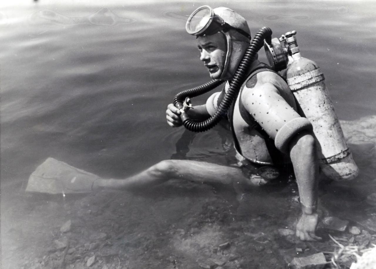 Historische schwarz-weiß Fotografie eines jungen Tauchers der am Ufer sitzt und zur Seite blickt. Zu seiner Ausrüstung gehören Schwimmflossen, eine Taucherbrille und eine Sauerstofflasche am Rücken. Historische schwarz-weiß Fotografie eines jungen Tauchers der am Ufer sitzt und zur Seite blickt. Zu seiner Ausrüstung gehören Schwimmflossen, eine Taucherbrille und eine Sauerstofflasche am Rücken.