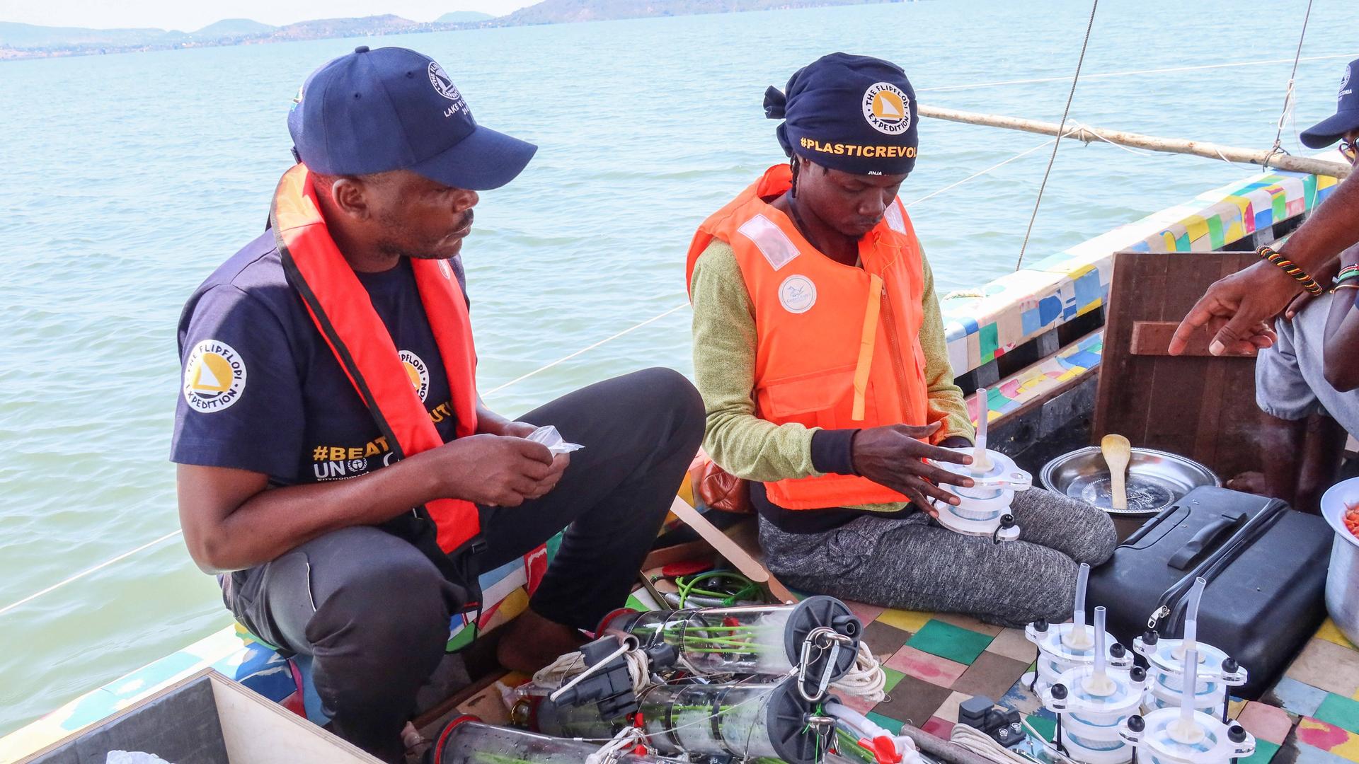 Zwei Männer sitzen auf einem Boot, vor ihnen sind Plastikdosen und -behälter, mit denen Wasserproben aus dem Victoriasee genommen werden. Der rechte Mann ist Kenias Superstar Juliani.