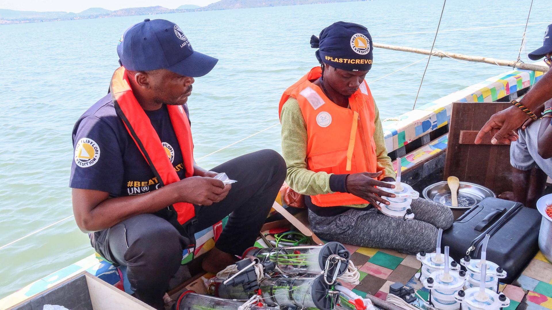 Zwei Männer sitzen auf einem Boot, vor ihnen sind Plastikdosen und -behälter, mit denen Wasserproben aus dem Victoriasee genommen werden. Der rechte Mann ist Kenias Superstar Juliani. Zwei Männer sitzen auf einem Boot, vor ihnen sind Plastikdosen und -behälter, mit denen Wasserproben aus dem Victoriasee genommen werden. Der rechte Mann ist Kenias Superstar Juliani.