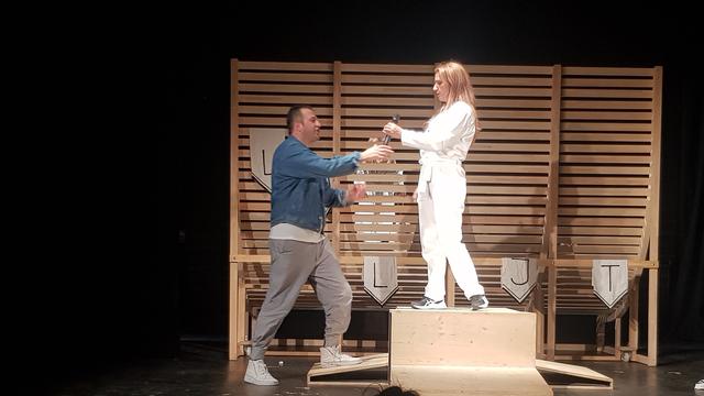 Auf einer Theaterbühne reicht ein Schauspieler seiner Kollegin ein Mikro. Er trägt einen blauen Pullover und eine weiße Hose. Sie steht auf einem Podest und ist ganz in weiß gekleidet.