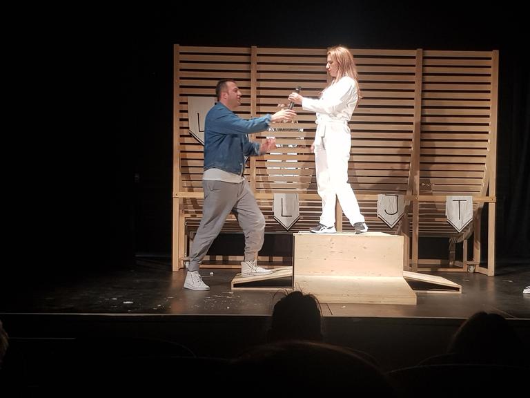 Auf einer Theaterbühne reicht ein Schauspieler seiner Kollegin ein Mikro. Er trägt einen blauen Pullover und eine weiße Hose. Sie steht auf einem Podest und ist ganz in weiß gekleidet.