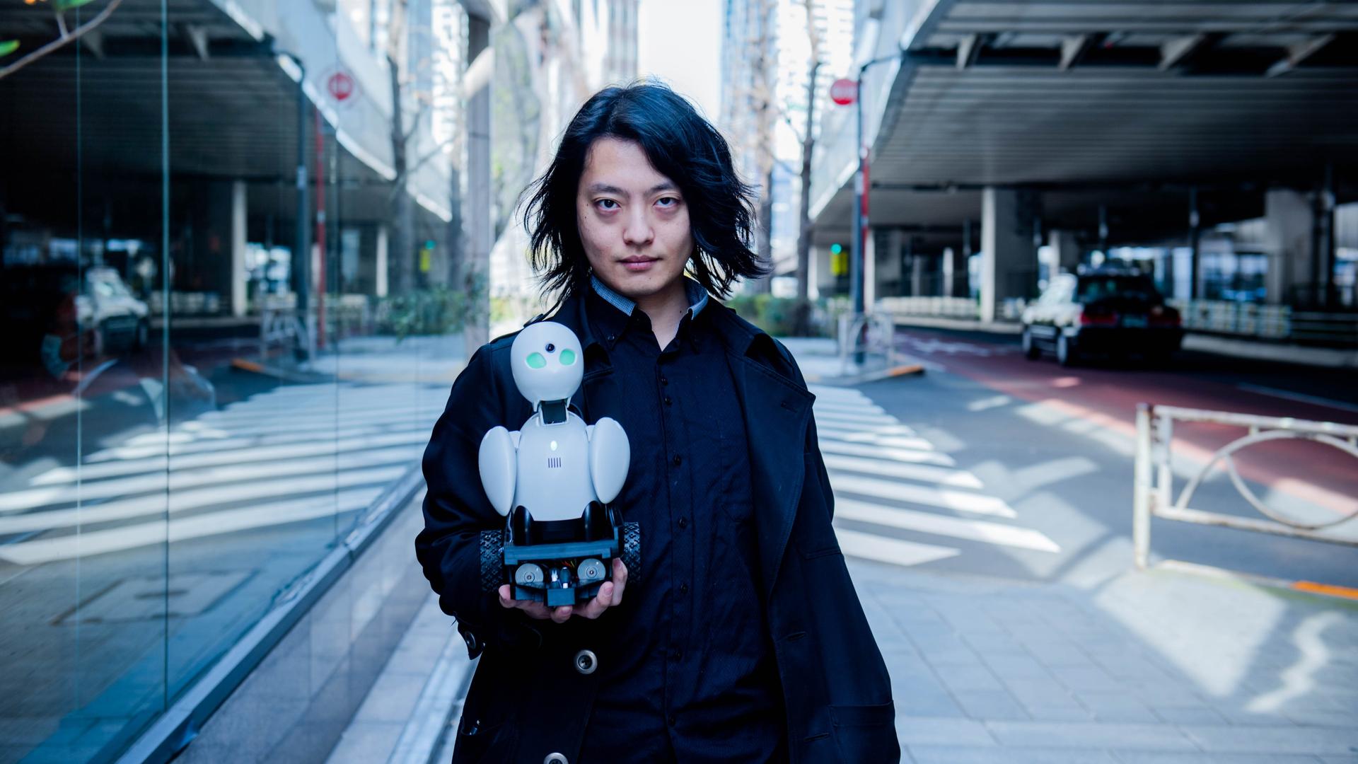 Der japanische Medienkünstler Ory Yoshifuji trägt ein weites schwarzes Sakko und hält in seiner rechten Hand einen 20 cm hohen weißen Roboter mit grünen Augen. Der japanische Medienkünstler Ory Yoshifuji trägt ein weites schwarzes Sakko und hält in seiner rechten Hand einen 20 cm hohen weißen Roboter mit grünen Augen.