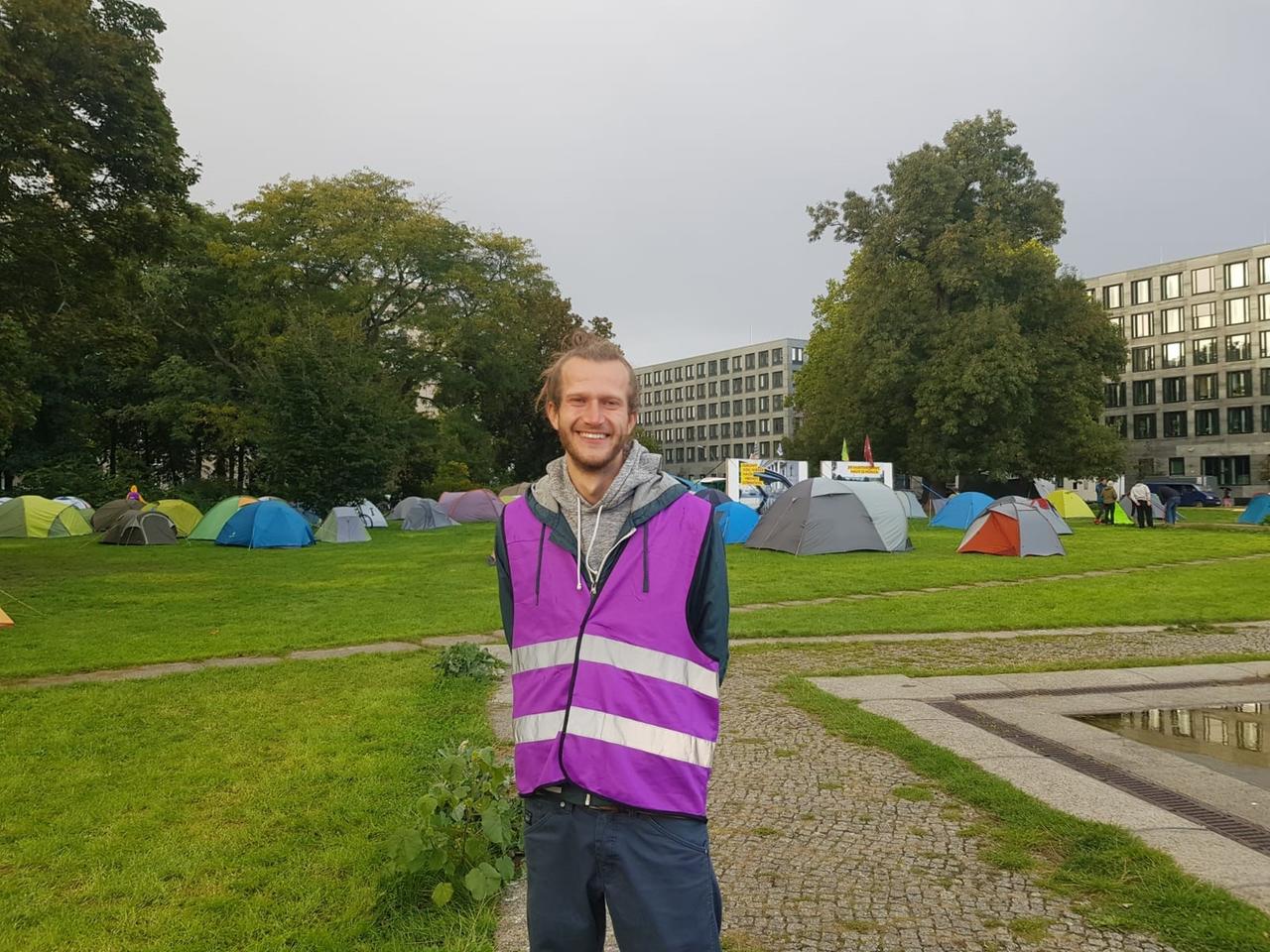 Aktivist Jan Gerber mit lila Sicherheitsweste, im Hintergrund ist ein Protestcamp zu sehen. Aktivist Jan Gerber mit lila Sicherheitsweste, im Hintergrund ist ein Protestcamp zu sehen.