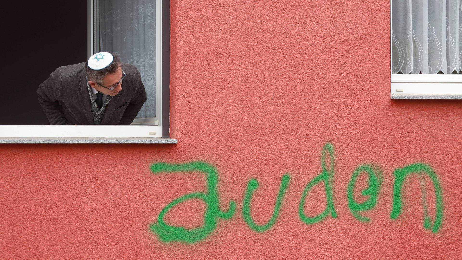 Ein Mann mit Kippa guckt aus einem Fenster. Unter dem Fenster ist die Wand mit dem Wort "Juden" beschmiert.