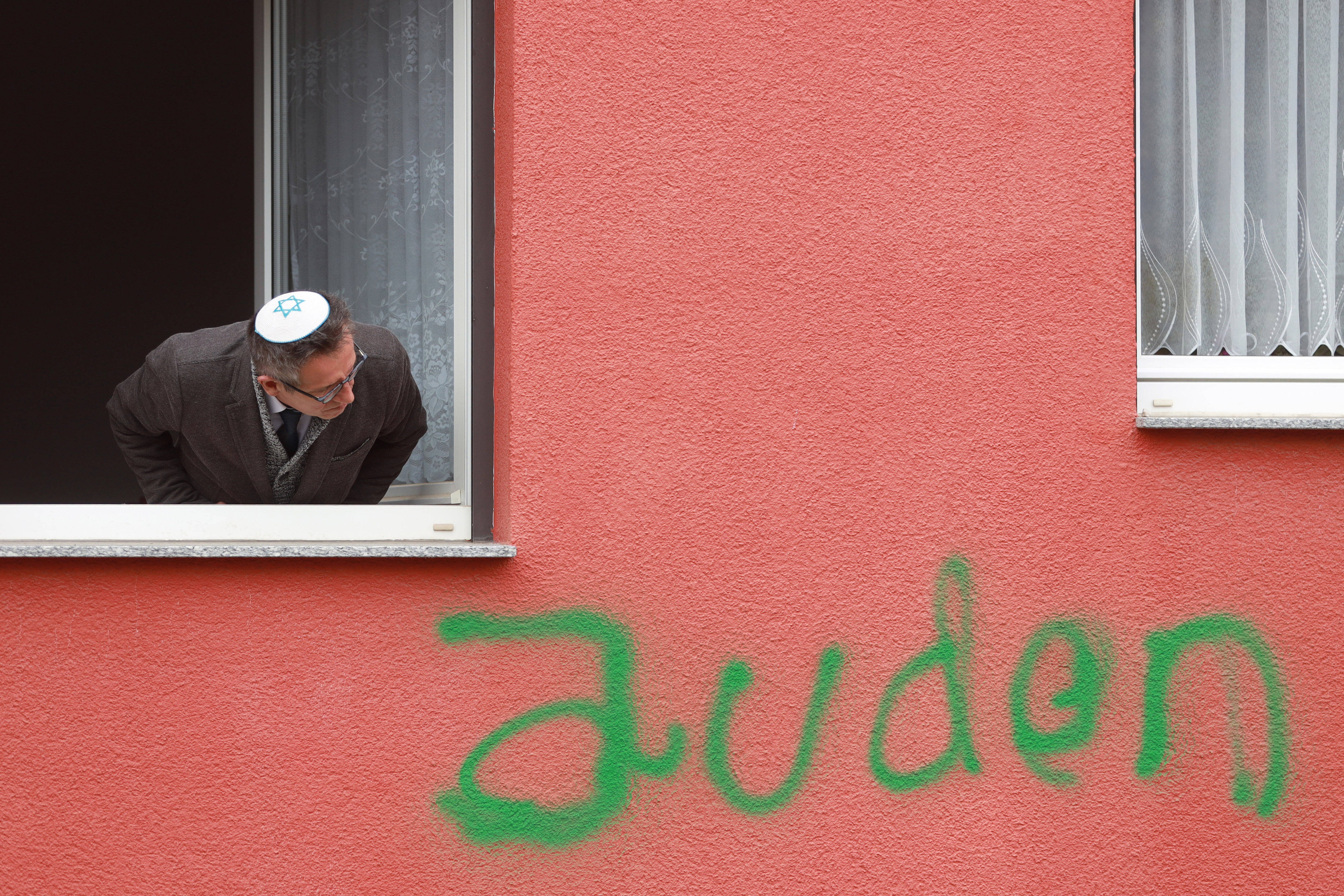 Ein Mann mit Kippa guckt aus einem Fenster. Unter dem Fenster ist die Wand mit dem Wort "Juden" beschmiert.
