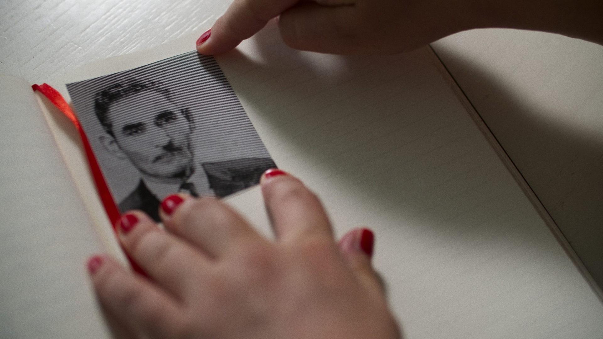 Hände mit rot lackierten Fingernägeln halten das Schwarz-Weiß-Bild eines Mannes mit Schnurrbart, Schlips und dunklem Sakko auf einer Albumseite. Hände mit rot lackierten Fingernägeln halten das Schwarz-Weiß-Bild eines Mannes mit Schnurrbart, Schlips und dunklem Sakko auf einer Albumseite.
