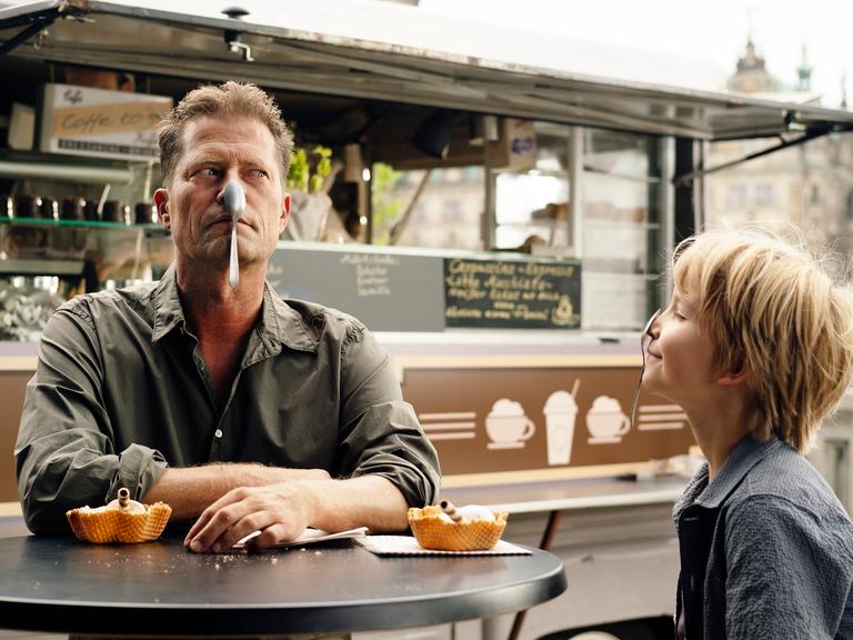 Filmszene aus dem Film "Lieber Kurt." Der Regisseur und Darsteller Til Schweiger sitzt mit einem Löffel im Gesicht an einem Tisch zusammen mit einem kleinen Jungen, der ebenfalls einen Löffel auf der Nase hat.