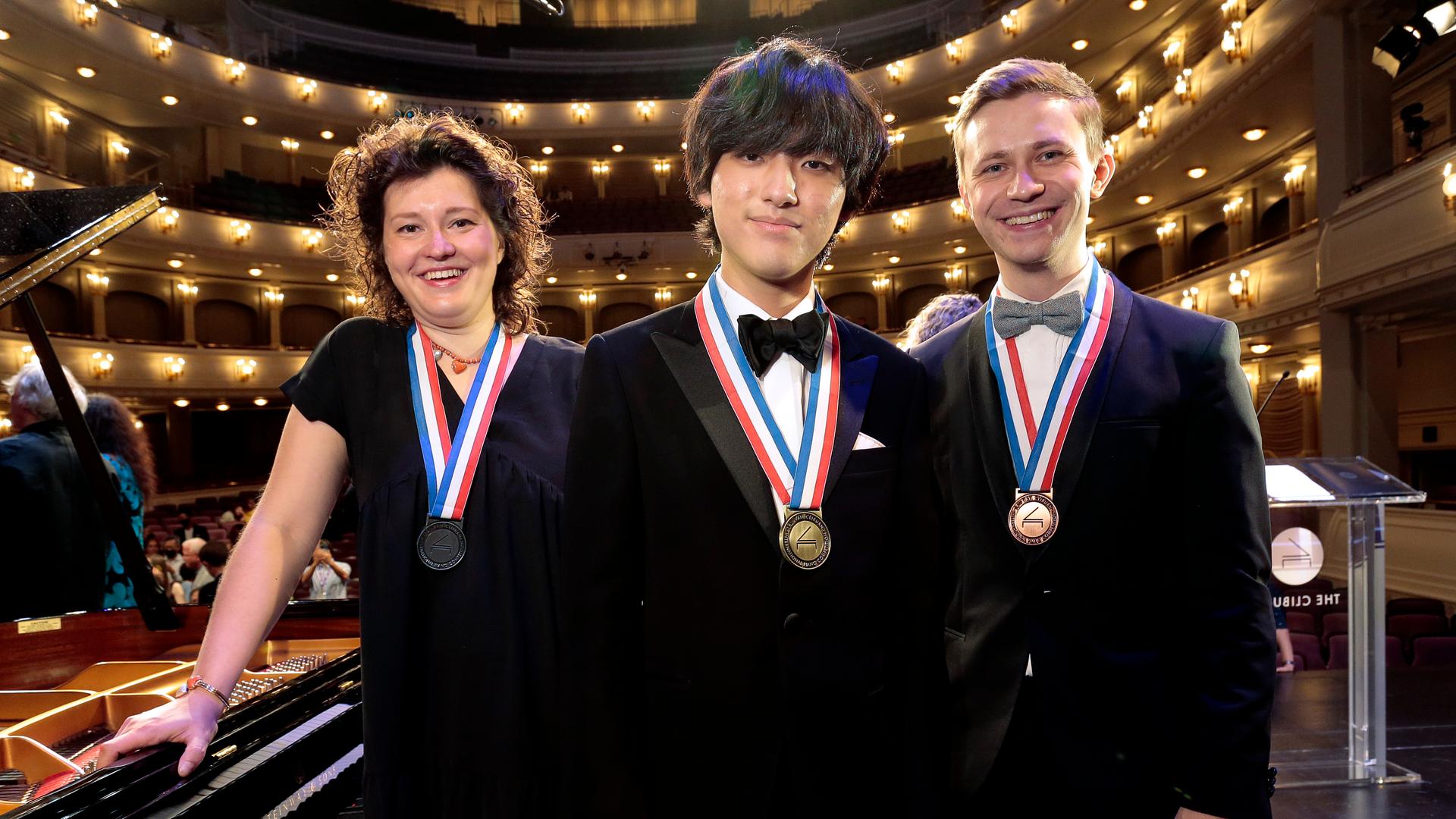 Eine Frau und zwei Männer stehen lachend mit Medaillen um den Hals neben einem Flügel. Im Hintergrund erkennt man einen Konzertsaal mit vielen Rängen.
