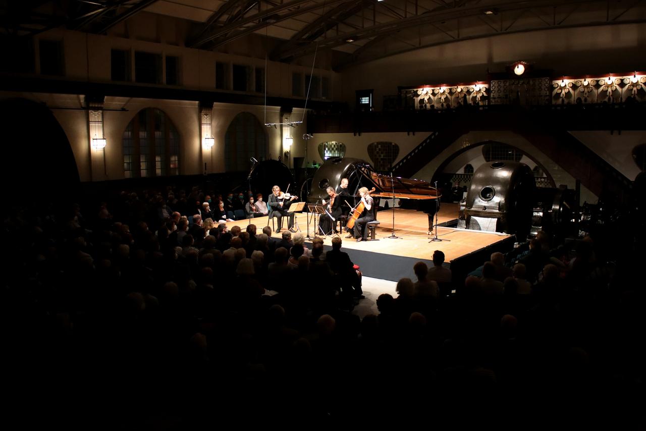 Ein Geiger, eine Bratschistin, ein Pianist und eine Cellistin musizieren auf einer Bühne. Umrahmt werden sie von Maschinen im Jugendstil-Kraftwerk Heimbach (Eifel) sowie Zuhörerinnen und Zuhörern.