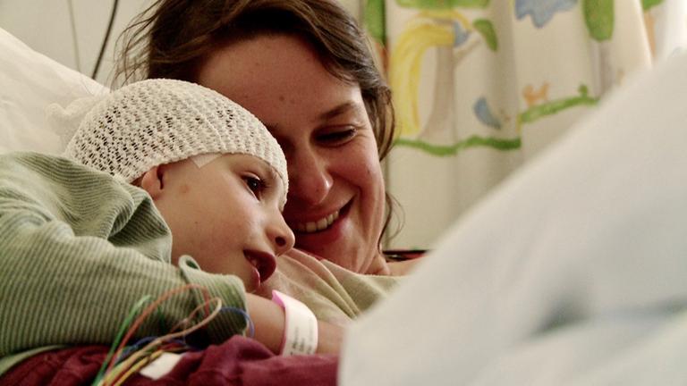 Eine junge Mutter liegt in einem Krankenhausbett und lächelt ihr Kind an, das sich an ihre Schulter schmiegt. Das Kind trägt ein Haarnetz, das ein Pflaster an der Schläfe halb verdeckt. Eine junge Mutter liegt in einem Krankenhausbett und lächelt ihr Kind an, das sich an ihre Schulter schmiegt. Das Kind trägt ein Haarnetz, das ein Pflaster an der Schläfe halb verdeckt.