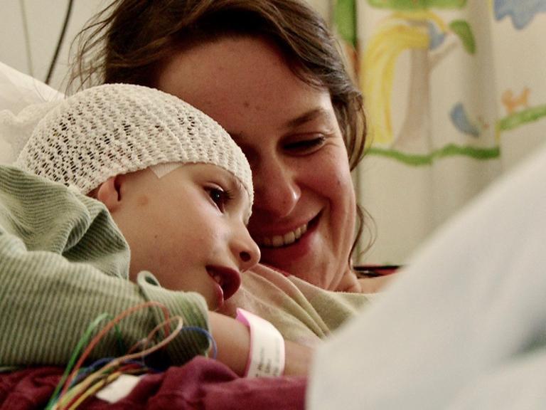 Eine junge Mutter liegt in einem Krankenhausbett und lächelt ihr Kind an, das sich an ihre Schulter schmiegt. Das Kind trägt ein Haarnetz, das ein Pflaster an der Schläfe halb verdeckt.