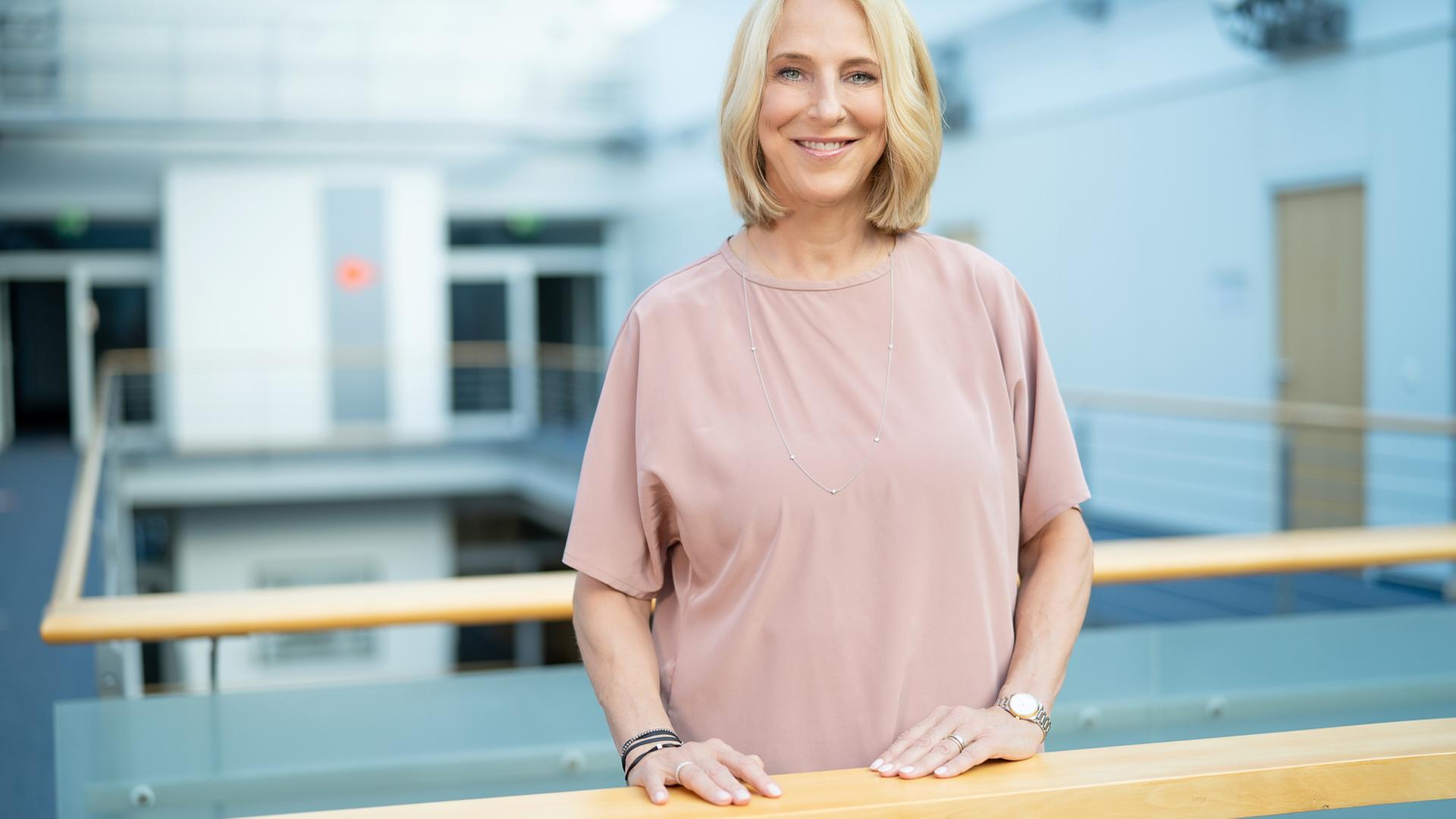 Eine blonde Frau steht in einem Bürogebäude und lächelt in die Kamera