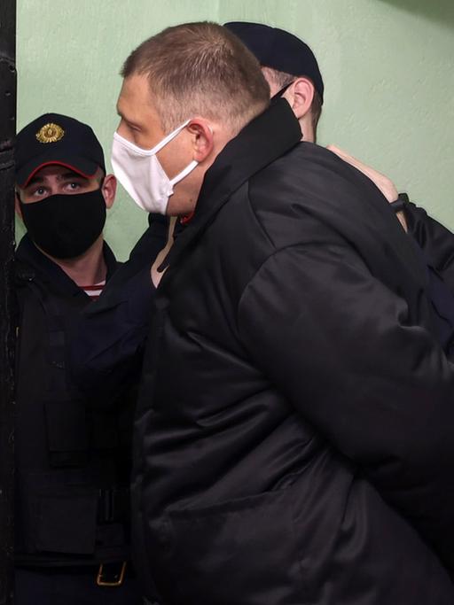 Ein kurzhaariger Mann in dunkler Jacke und mit Mund-Nasenschutz wird von Polizisten abgeführt.