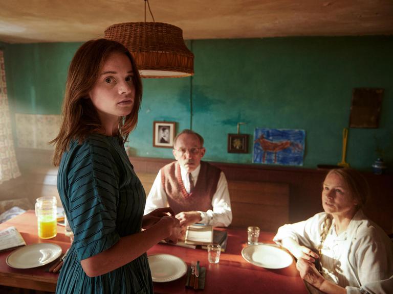 Im Filmstill aus "Was man von hier aus sehen kann" steht Luna Wedler im grünen Kleid in einer Küche und schaut direkt in die Kamera. Hinter ihr am Esstisch sitzen ihre Großeltern und schauen zu ihr auf.