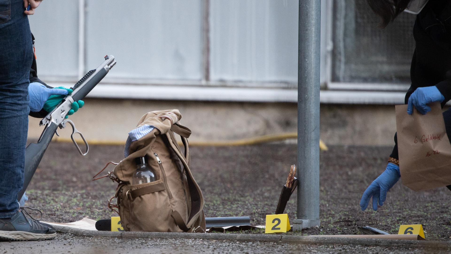 Nahaufnahme einer Spurensicherung: Zwei Personen mit blauen Schutzhandschuhen hocken auf dem Boden vor einem Gebäude und untersuchen ein Gewehr und einen Rucksack. Nahaufnahme einer Spurensicherung: Zwei Personen mit blauen Schutzhandschuhen hocken auf dem Boden vor einem Gebäude und untersuchen ein Gewehr und einen Rucksack.