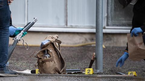 Nahaufnahme einer Spurensicherung: Zwei Personen mit blauen Schutzhandschuhen hocken auf dem Boden vor einem Gebäude und untersuchen ein Gewehr und einen Rucksack.