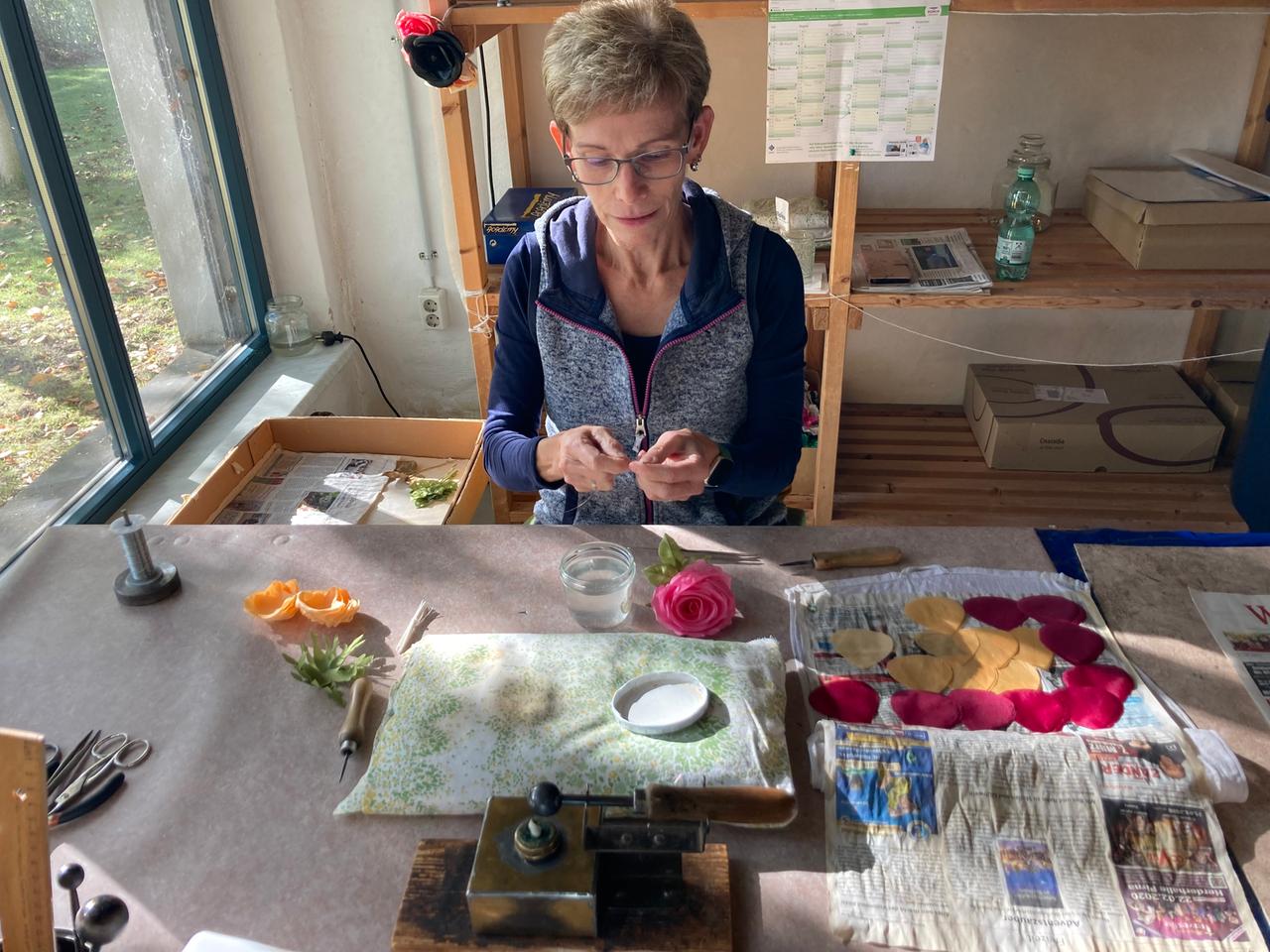 Eine Mitarbeiterin der Kunstblumenmanufaktur Heide Steyer sitzt an ihrem Arbeitstisch und fertigt Blüten aus Stoff. Vor ihr auf dem Tisch liegt ein Steckkissen, daneben Blütenblätter aus Stoff sowie bereits fertiggestellte Blumen. Die Frau trägt eine Brille und hat kurze graue Haare. Eine Mitarbeiterin der Kunstblumenmanufaktur Heide Steyer sitzt an ihrem Arbeitstisch und fertigt Blüten aus Stoff. Vor ihr auf dem Tisch liegt ein Steckkissen, daneben Blütenblätter aus Stoff sowie bereits fertiggestellte Blumen. Die Frau trägt eine Brille und hat kurze graue Haare.