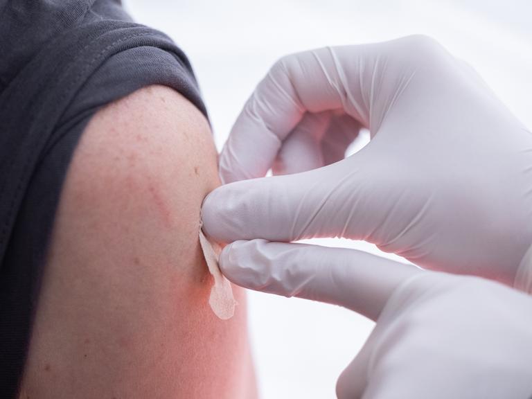 Eine geimpfte Person bekommt vom Arzt ein Pflaster auf die Nadeleinstichstelle am Oberarm.