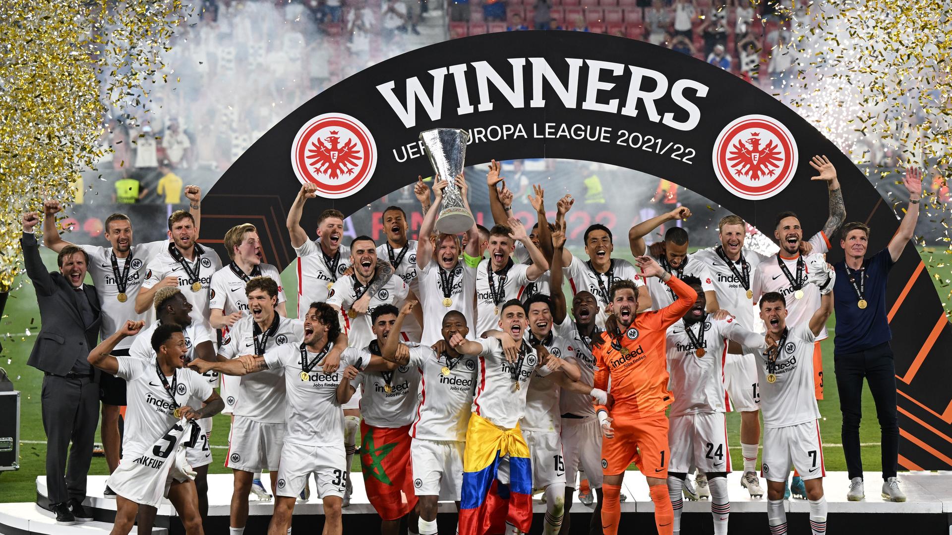 Die Spieler von Eintracht Frankfurt jubeln über den Gewinn  der Europa League. Sie stehen unter einem Bogen, auf dem groß geschrieben "Winners - UEFA Europa Legue 2021/22" zu lesen ist. Einer der Spieler reckt den Pokal in die Höhe.