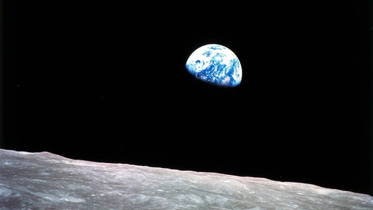 Beim Blick über eine steinige Ebene des Mondes erleuchtet die blau-weiß schimmernde Erde zur Hälfte am stockfinsteren Himmel. 