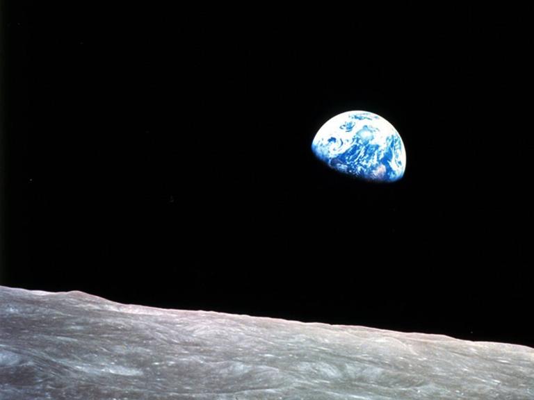 Beim Blick über eine steinige Ebene des Mondes erleuchtet die blau-weiß schimmernde Erde zur Hälfte am stockfinsteren Himmel. 