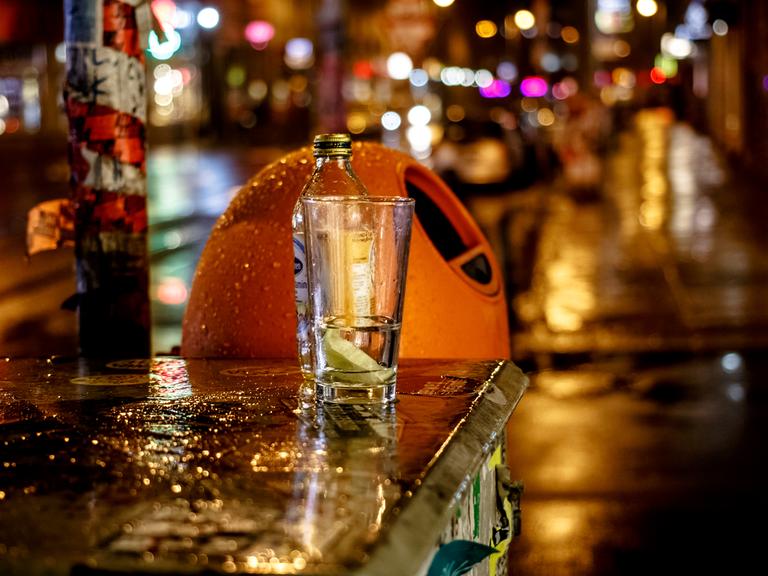 Nächtliche Straßenszene bei Regenwetter in Berlin. Zu sehen ist ein typischer orangefarbener Mülleimer. Davor, auf einem Kasten, steht eine Flasche und ein Glas. 