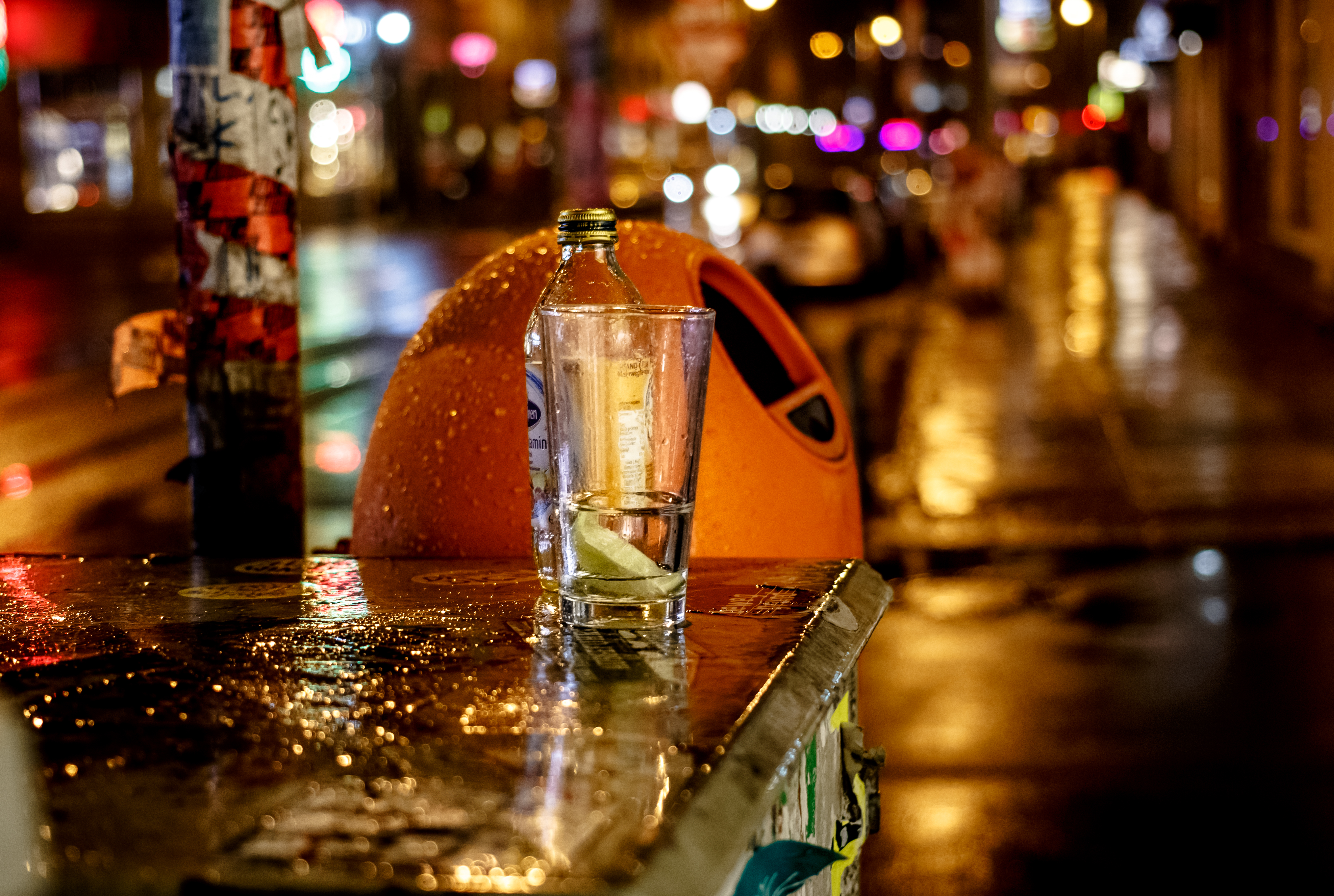 Nächtliche Straßenszene bei Regenwetter in Berlin. Zu sehen ist ein typischer orangefarbener Mülleimer. Davor, auf einem Kasten, steht eine Flasche und ein Glas. 
