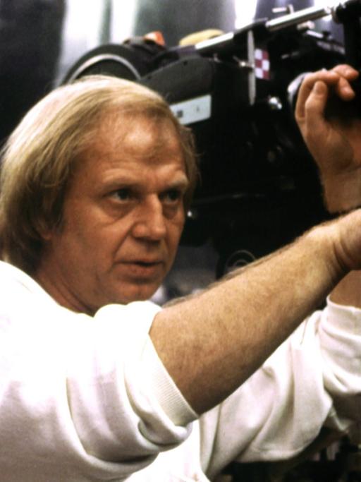 Der Regisseur Wolfgang Petersen, neben einer großen Filmkamera, gibt Handzeichen in Richtung der Schauspieler.