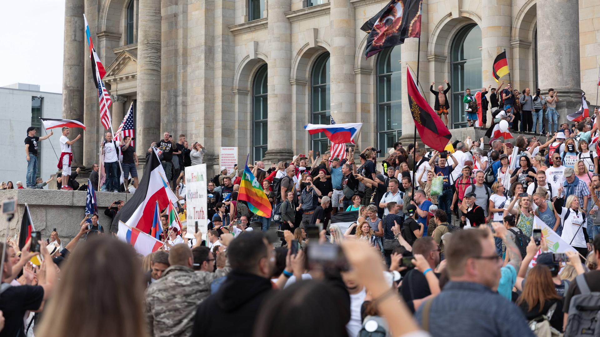 Blick auf den Reichstag, vor dem Hunderte Demonstrierende gegen den Corona-Maasnahmen stehen. Dabei stürmen einige die Treppen des Reichstages. Eine Vielzahl von Flaggen ist zu sehen. Unter anderen die Regenbogenflagge, die Flagge der USA, die russische und die deutsche. Blick auf den Reichstag, vor dem Hunderte Demonstrierende gegen den Corona-Maasnahmen stehen. Dabei stürmen einige die Treppen des Reichstages. Eine Vielzahl von Flaggen ist zu sehen. Unter anderen die Regenbogenflagge, die Flagge der USA, die russische und die deutsche.