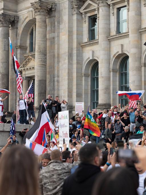 Blick auf den Reichstag, vor dem Hunderte Demonstrierende  gegen den Corona-Maasnahmen stehen. Dabei stürmen einige die Treppen des Reichstages. Eine Vielzahl von Flaggen ist zu sehen. Unter anderen die Regenbogenflagge, die Flagge der USA, die russische und die deutsche.