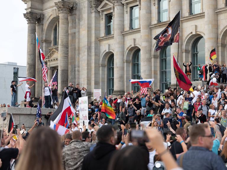 Blick auf den Reichstag, vor dem Hunderte Demonstrierende  gegen den Corona-Maasnahmen stehen. Dabei stürmen einige die Treppen des Reichstages. Eine Vielzahl von Flaggen ist zu sehen. Unter anderen die Regenbogenflagge, die Flagge der USA, die russische und die deutsche.