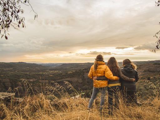 Drei junge Frauen sind mit dem Rücken zur Kamera zu sehen und stehen in der Landschaft. 