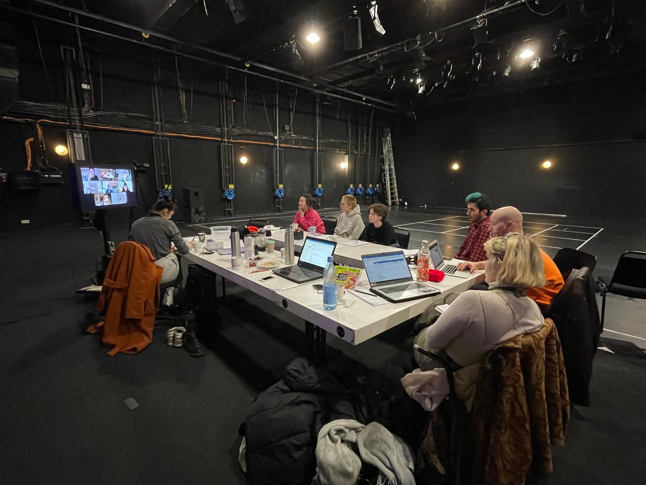 In einem dunklen Raum, der der Hintergrund einer Theaterbühne zu sein scheint, steht ein großer Tisch an dem mehrere Personen sitzen. Sie schauen auf einen großen Bildschirm auf dem eine Videokonferenz mit weiteren Personen stattfindet. In einem dunklen Raum, der der Hintergrund einer Theaterbühne zu sein scheint, steht ein großer Tisch an dem mehrere Personen sitzen. Sie schauen auf einen großen Bildschirm auf dem eine Videokonferenz mit weiteren Personen stattfindet.