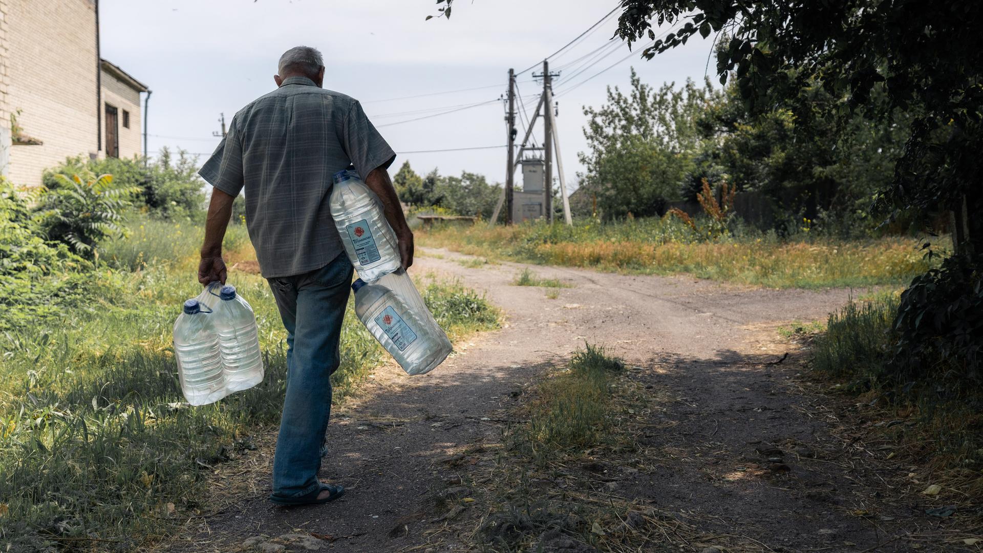 Zu sehen ist ein Mann, der große Wasserflaschen in den Händen und unter den Armen trägt. Er geht einen unbefestigten Weg entlang, der zwischen Strommasten und einem Gebäude verläuft.