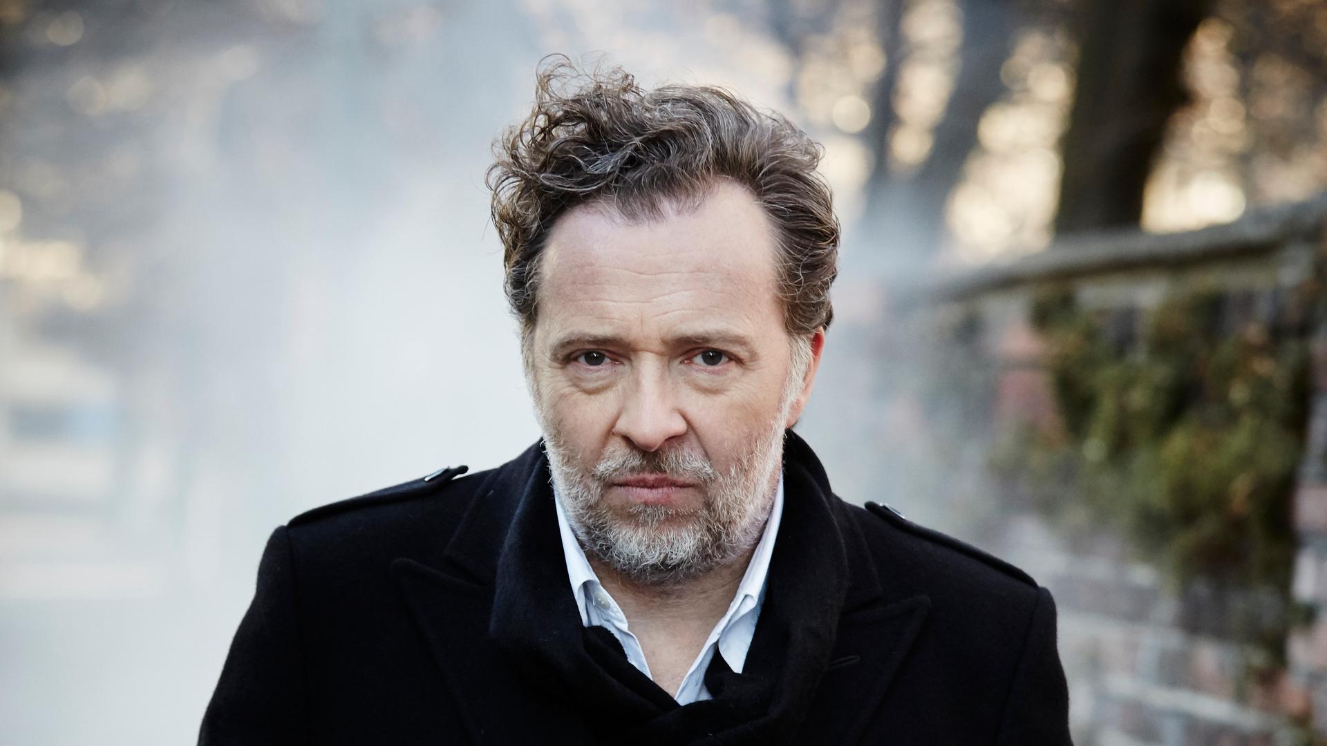 Christian Gerhaher steht im Freien. Er hat kurze, lockige Haare, trägt einen Bart und einen dunklen Mantel. Rechts ist eine Mauer zu erkennen. Christian Gerhaher steht im Freien. Er hat kurze, lockige Haare, trägt einen Bart und einen dunklen Mantel. Rechts ist eine Mauer zu erkennen.