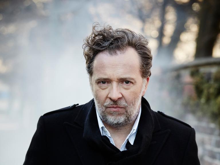 Christian Gerhaher steht im Freuen. Er hat kurze, lockige Haare, trägt einen Bart und einen dunklen Mantel. Rechts ist eine Mauer zu erkennen.