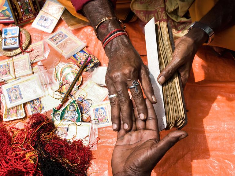Ein Wahrsager hält die Hand seines Kunden. Daneben liegen Karten mit den Bildern hinduistischer Götter und ein Gewirr roter Schnüre.