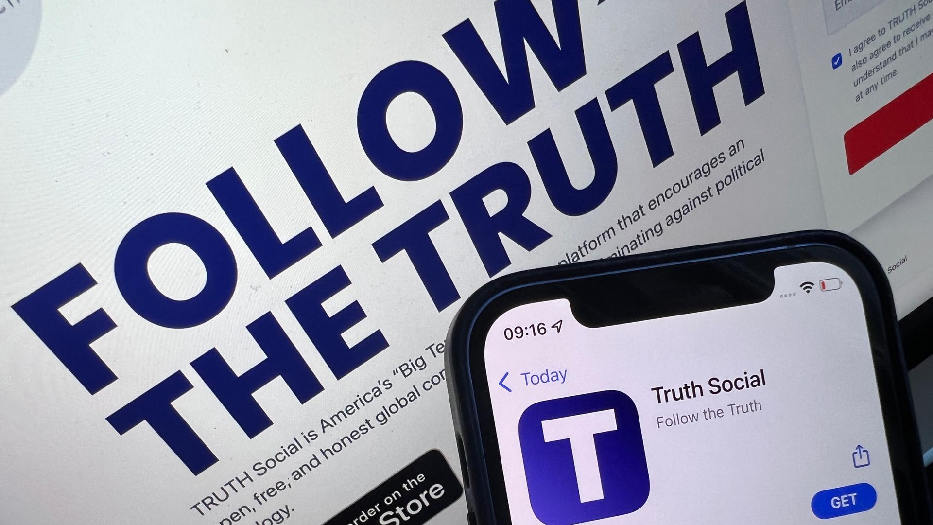 Das Bild zeigt ein Handy, auf dem die "Truth Social"-Anwendung geöffnet ist. Dahinter auf einem Bildschirm die Ankündigung des Starttermins.
