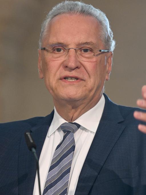 Der Bayerische Innenminister Joachim Herrmann spricht mit erhobener Hand vor zwei Mikrofonen.