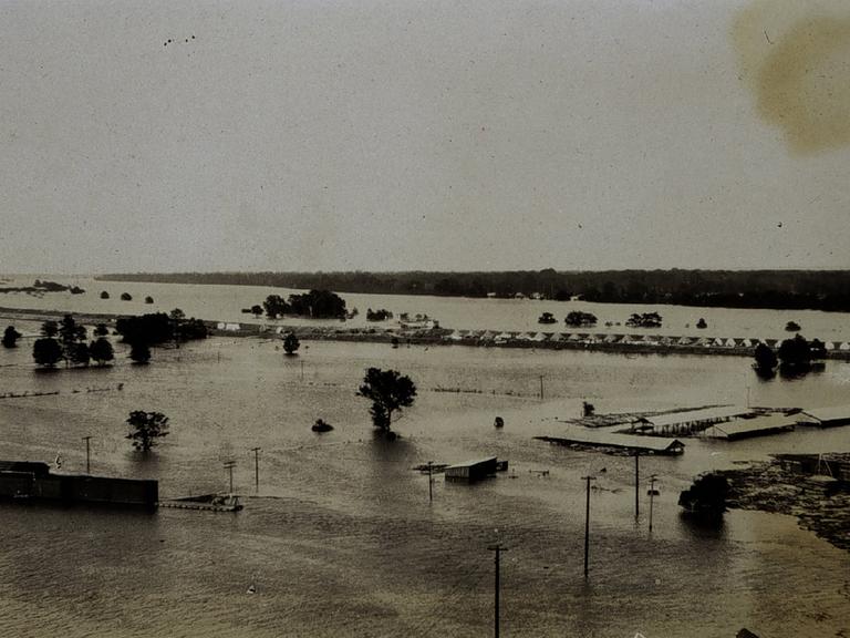 Die historische Schwarz-Weiß-Fotografie aus dem Jahr 1927 zeigt Wassermassen, aus denen vereinzelt Dächer eines Zeltcamps für Flüchtlinge herausragen. 