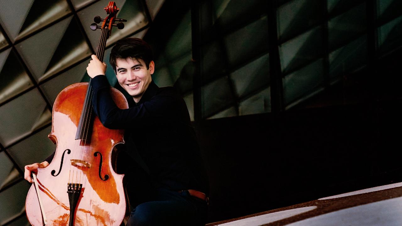 Ein junger Mann mit kurzen schwarzen Haaren lacht. Er hält ein Cello in der linken Hand. Hinter ihm erkennt man einen gemusterten Hintergrund.