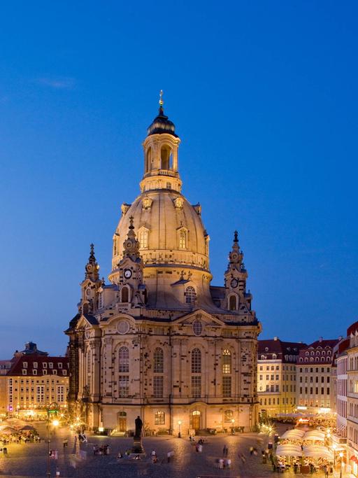 Blick auf den abendlichen, aber erleuchteten Neumarkt in Dresden auf die Frauenkirche vor blauem Himmel. 