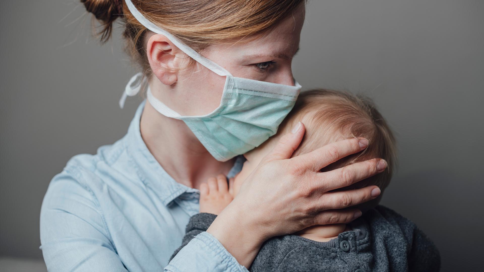 Eine Mutter mit medizinischer Maske trägt ihr krankes Kleinkind auf dem Arm. Eine Mutter mit medizinischer Maske trägt ihr krankes Kleinkind auf dem Arm.