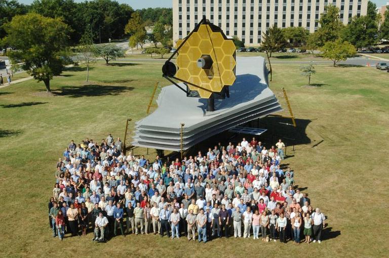 Mitarbeiterinnen und Mitarbeiter am JWST-Projekt stehen vor einem Modell des Weltraumteleskops