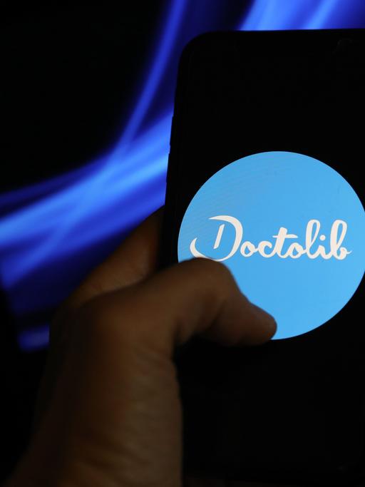 Eine Hand hält ein Smartphone, auf dem das Logo von Doctolib zu sehen ist. 