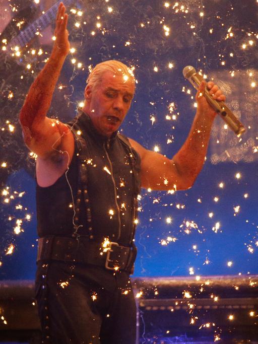 Till Lindemann steht mit erhobenen Händen auf einer Bühne, von goldenem Konfettiregen umgeben
