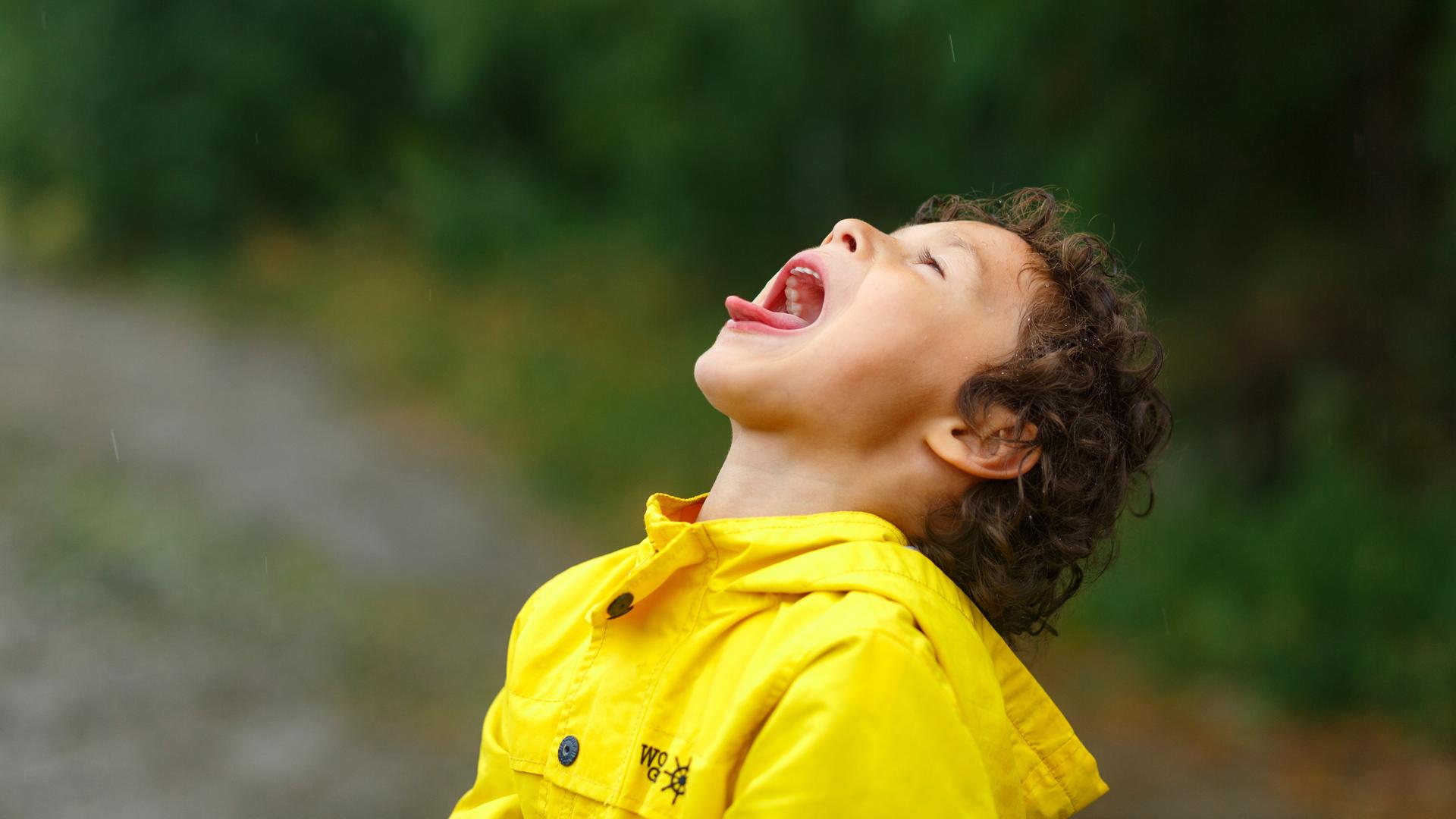 Ein Kind in gelber Regenjacke legt den Kopf in den Nacken und streckt mit weit geöffnetem Mund die Zunge heraus. Ein Kind in gelber Regenjacke legt den Kopf in den Nacken und streckt mit weit geöffnetem Mund die Zunge heraus.