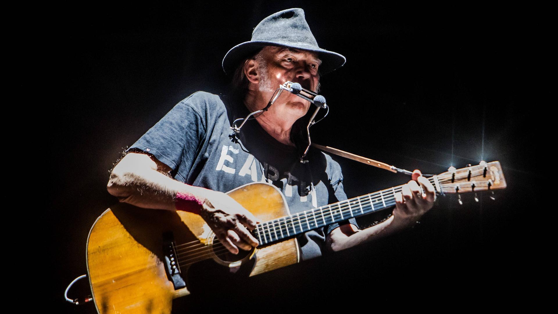 Der US-Musiker Neil Young steht mit Munharmonika und Gitarre auf einer Bühne. Er trägt einen Hut. 