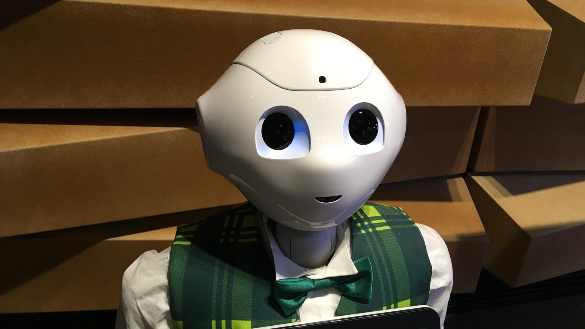 Kopf und Schultern des humanoiden Roboters "Pepper": glatte weiße Oberfläche, sehr große Augen, eine angedeutete Nase und ein kleiner lächelnder Mund. Er trägt eine karierte Weste und eine Fliege.